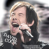 A person with long dark hair and light brown highlights sings into a black and silver microphone, wearing a black shirt with white text.
