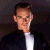 Pixelated portrait of a person with short dark hair wearing a dark jacket over a white shirt against a dark background.