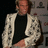 Pixelated image of a person wearing a black shirt and light-colored jacket with dark patterns, standing against a dark background.
