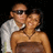 Two people in close proximity; one wears sunglasses and a white shirt, the other has dark hair and no visible clothing. Background is solid black.
