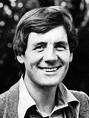 Black-and-white photograph of a smiling person with short dark hair wearing a collared shirt and textured jacket against a blurred outdoor background.