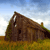 Pixelated static image of a brown wooden structure with a sloped roof, dark rectangular opening, and greenery on a grassy field under a blue sky.