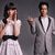 Two figures in formal attire stand against a gray background; the left figure covers their face with hands while the right figure holds a microphone.
