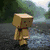 A pixelated Danbo character stands on a gravel path with greenery in the background, then shifts to holding a glowing sword-like object.