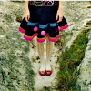 A person stands in shallow water wearing a dark skirt with blue and pink trim, white socks, and red shoes, with hands resting on the skirt.