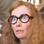 Pixelated close-up of a person with light brown hair, wearing dark-rimmed glasses, against a blurred background with brown and beige tones.