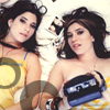 Two women in yellow and white patterned outfits lie side-by-side on a white surface, one holding a blue and black object with visible text.