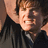 Pixelated close-up of a person with light brown hair wearing dark clothing, showing partial face and shoulder in warm-toned lighting.