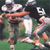 Pixelated static image of two football players in mid-action on a green field, one player wearing white jersey with number 19 and dark helmet, the other in orange uniform with light-colored helmet, both engaged in physical contact during a game.