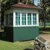 Pixelated image of a small green building with white trim around windows and a brown roof, set against a blurred background of trees and grass.