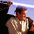 Pixelated static image of a character in brown and beige clothing holding a glowing white lightsaber against a dark background with purple sky elements.