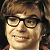 Pixelated close-up of a person with brown hair, black-framed glasses, and a slight smile, wearing a red garment against a dark background.