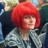 Static image of a person with bright red hair wearing a light gray jacket over dark clothing against a blurred background.