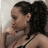 A woman with dark curly hair in a high ponytail, wearing a black spaghetti-strap top, resting her chin on her hand against a light background.