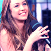 A person with long brown hair holds a black microphone with a teal stripe, smiling with teeth visible against a blurred background.