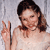 A person with light brown hair tied back in a ponytail, wearing a light-colored sleeveless top, making a peace sign with their right hand against a light gray background.
