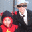 Two figures in a pixelated image: one wearing red with yellow symbol, the other in dark jacket and white shirt. Background shows blurred outdoor scene with vertical pole.