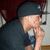 Pixelated image of a person wearing dark clothing and a cap, holding an object near their face against a blurred background.