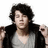 A person with dark curly hair wears a gray V-neck shirt and black jacket, holding the jacket open with both hands against a white background.