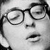 Black-and-white pixelated image of a person's face wearing round eyeglasses, with visible eyes, nose, mouth, and hair.