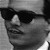 Black-and-white pixelated image of a person wearing dark sunglasses and a light-colored cap, cropped to show the upper face and shoulders.