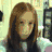 A static image of a person with long brown hair in a light-colored room, wearing dark clothing and holding an object near their face.