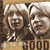 Two figures with light brown hair smile beneath a sign reading 'GOOD' while another figure in dark clothing holds a wand-like object against a muted background.