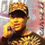 A person wearing a camouflage cap and dark sweater with gold patterns holds a phone to their ear against a pinkish background.