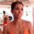 A person in an orange bikini top stands in the foreground with a blurred background showing a pool area and another figure in swimwear.