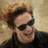 A person with light brown curly hair and dark sunglasses wears a black turtleneck, smiling slightly against a beige textured background.