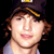 Static pixelated image of a person wearing a dark cap with yellow trim and light-colored shirt against black background.