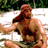 Pixelated static image of a shirtless man with long brown hair and beard wearing camouflage shorts, standing outdoors on a white surface with green foliage background.