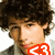 A pixelated image of a person with curly dark hair, wearing a light-colored shirt and gray jacket, with red text overlay on the lower left.