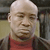 Pixelated close-up of a person with a bald head wearing a red shirt, set against a blurred background with indistinct shapes and muted colors.