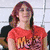 A person with red hair wears a red graphic t-shirt and black pants, seated with arms crossed, with a green garment and black text reading 'dul' visible below.