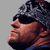 Pixelated static image of a person's face in profile view with dark hair and a fur-lined hood against a gray background.