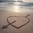 A beach scene with a heart-and-arrow drawing in sand, featuring text overlays that change across frames.