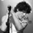 Black-and-white pixelated image of a person with dark hair holding a microphone stand, smiling slightly against a gradient background.
