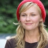 A person with long blonde hair wears a red knit beret and a dark brown top against a blurred green background.