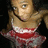 Pixelated image of a person with light skin wearing red and white clothing, dark hair tied back, and a black strap across the chest against a dark background.