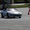 White convertible sports car in motion on a paved road with orange traffic cones and people standing nearby.
