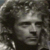 Pixelated grayscale image of a human face with dark hair, neutral expression, and visible eyes, nose, and mouth against a dark background.