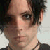 Pixelated close-up of a person with dark tousled hair, light skin, and a neutral expression, wearing a dark top with a thin necklace.