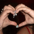 Two hands form a heart shape with fingers, wearing rings on multiple fingers, against a dark background with a red line visible at the bottom right.