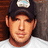 A pixelated close-up of a person wearing a dark cap with a light-colored emblem, light skin, and a white shirt against a brown and gray background.