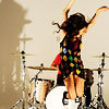 A person with dark hair plays drums, arms raised, wearing a colorful patterned dress with red, green, yellow, and blue shapes on a dark background.