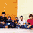 Pixelated static image of five people seated in a row against an orange and beige background with scattered white dots.