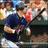 A baseball player in a dark blue jersey and white pants swings a bat, with another player in red visible behind him.
