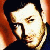 Pixelated static image of a person's face with dark hair and light skin tones, rendered in warm orange-brown hues against a black background.