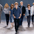 Group of people walking in a line, with a central figure in blue shirt and dark pants leading the group against a gray background.